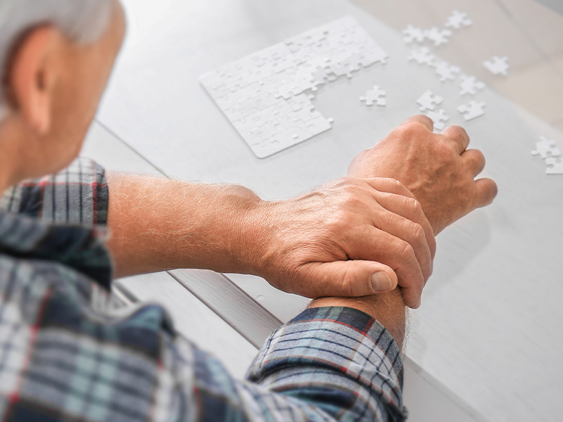A imagem mostra um homem branco, de cabelos grisalhos, no canto esquerdo da imagem. Ele está de costas, e é possível visualizar seu ombro direito e seus braços, sobre uma mesa branca. Ele veste camisa xadrez em tons de azul e vermelho, e com a mão direita monta um quebra-cabeças que está sob a mesa. Com a mão esquerda, ele segura seu punho direito para controlar os movimentos involuntários da Doença de Parkinson. 